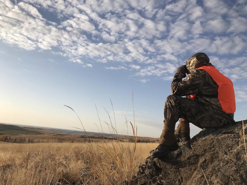 A hunter sitting on a rock in a field, using binoculars to look in the distance