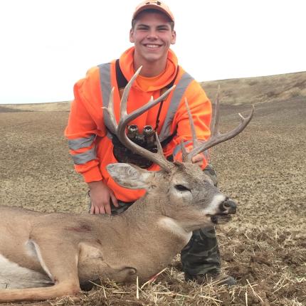 Young hunter wearing hunter orange jacket and binoculars sitting next to harvested white tailed deer.