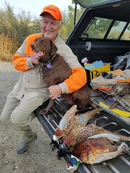 A hunter sitting on the tailgate of his care hugging his dog with three freshly harvested pheasants