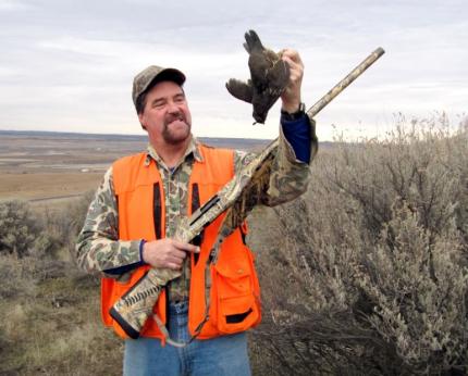 A hunter wearing a hunter orange vest holding a shotgun and a freshly harvested quail.